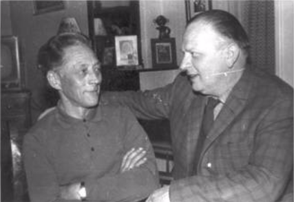 Two men are sitting indoors. One man has his arms crossed and is looking at the other, who is speaking with his arm resting on the first man's shoulder. Household items are visible in the background.