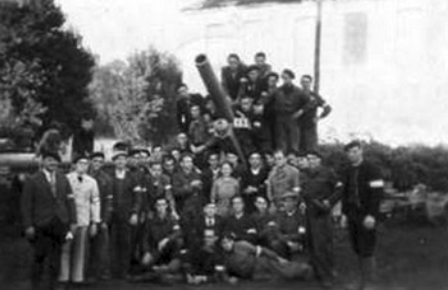 A large group of people, some in uniform, pose in front of a large artillery cannon outdoors with trees and a building in the background.