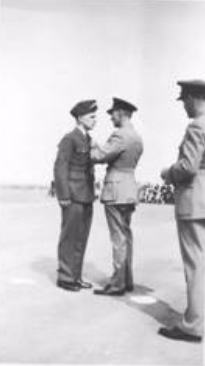 A military officer pins a medal or insignia on another uniformed man whilst a third man stands nearby on an open parade ground.