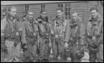 Six men in military uniforms and flying gear stand together outside a wooden building, posing for a group photo.