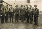 A group of uniformed airmen stand in front of a military aeroplane, posing for a photo on an airfield.