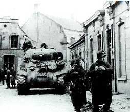 Black and white photo of soldiers walking in front of a tank on a narrow street lined with buildings, likely from an historical wartime setting.