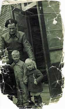 A man in a uniform stands behind two young children in coats, posing outside a building; the photo is old and creased.