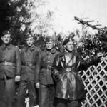 Four men in military uniforms stand outdoors near a lattice fence and foliage; one wears a long leather coat and points to the side. The photo is black and white.