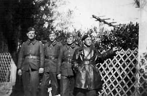 Four men in military uniforms stand outdoors near a lattice fence and foliage; one wears a long leather coat and points to the side. The photo is black and white.