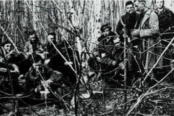 A group of armed men in coats and uniforms sit and stand among sparse trees and branches in a forested area.