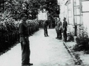 A group of soldiers stands in formation outdoors, facing a few individuals near a building with open windows and trees in the background.