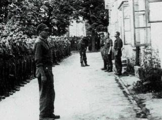 A group of soldiers stands in formation outdoors, facing a few individuals near a building with open windows and trees in the background.