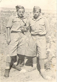 Two men in military uniforms and shorts stand side by side outdoors, posing for a photo on rocky terrain with sparse vegetation in the background.