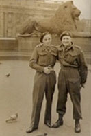Two people in military uniforms stand arm in arm in front of a large lion statue in a public square, with pigeons on the ground nearby.