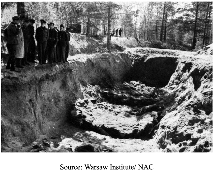 A group of people stands at the edge of a large mass grave in a forested area; partially buried bodies are visible in the pit.