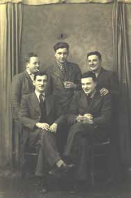 Five men in suits pose together for a formal, vintage black-and-white portrait in front of a plain curtain backcloth.