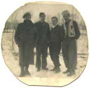 Four men stand together outdoors in winter, wearing coats and scarves, with snow on the ground and bare trees in the background.
