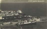 A large ship is docked at a port beside a coastal town with several buildings visible near the shoreline.