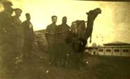 Five people stand next to a camel in an outdoor area with buildings in the background; the image appears old and sepia-toned.