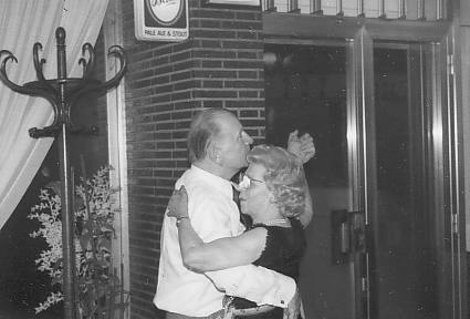 An older couple dances closely together indoors near a glass door and a coat rack, with a “no parking” sign visible on the wall behind them.