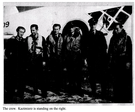 Six men in flight gear stand in front of an aeroplane; one man on the far right wears a dark uniform and hat.
