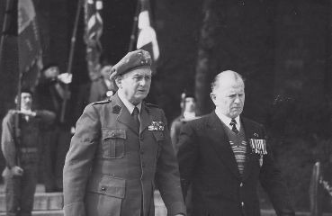Two men in military attire and suits with medals walk side by side, with uniformed individuals holding flags in the background.