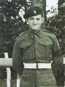 A young man in a military uniform and beret stands outdoors, facing the camera with his hands behind his back. Trees and a fence are visible in the background.