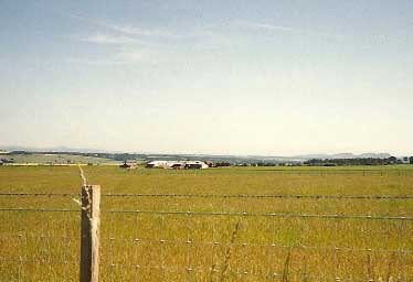 A barbed wire fence in the foreground with a wide grassy field and distant farm buildings under a clear sky.