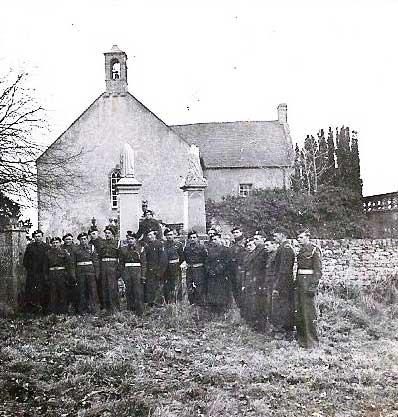 A group of uniformed men stand in front of a small church with statues and a bell tower, set in a grassy area.