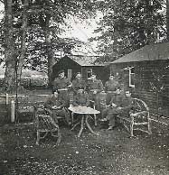 A group of soldiers in uniform pose around a table and chairs outdoors, with trees and wooden buildings in the background.