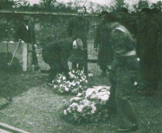 A group of people stands near graves adorned with floral arrangements during a burial ceremony in an outdoor setting.