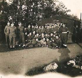 A group of uniformed soldiers pose together in front of a building with trees in the background.