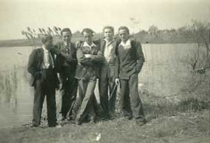Five men in suits stand together by the edge of a lake with reeds and trees in the background on a cloudy day.