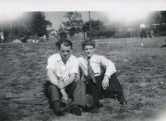 Two young men sit on the grass in an open field, both wearing collared shirts and trousers; trees, buildings, and other people are visible in the background.