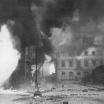 A city street with buildings in ruins and heavy smoke rising, likely due to a recent bombing or fire during wartime.