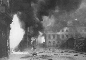 A city street with buildings in ruins and heavy smoke rising, likely due to a recent bombing or fire during wartime.