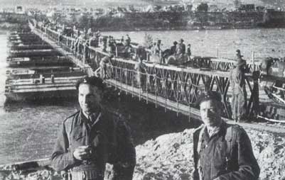 Two soldiers stand in the foreground while troops and vehicles cross a temporary military bridge over a river in a wartime setting.