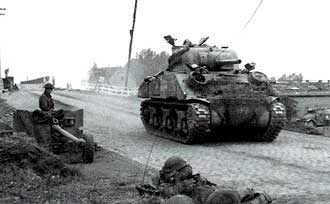 A World War II-era tank drives down a cobblestone road as soldiers, some manning artillery, are positioned along the roadside.
