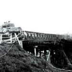 A military tank crosses a wooden bridge supported by beams, with grassy ground in the foreground and a body of water below the bridge.