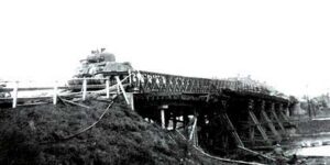 A military tank crosses a wooden bridge supported by beams, with grassy ground in the foreground and a body of water below the bridge.