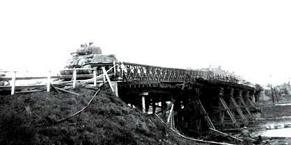 A military tank crosses a wooden bridge supported by beams, with grassy ground in the foreground and a body of water below the bridge.