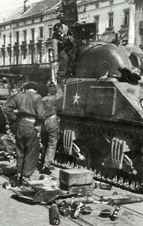 Three soldiers stand next to and on top of a Second World War-era tank, which is surrounded by equipment and supplies in an urban setting.
