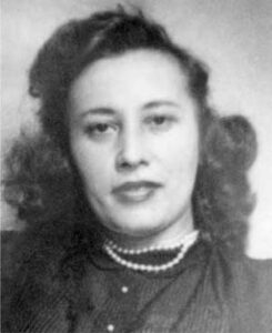 Black and white portrait of a woman with wavy hair, wearing a dark top and a pearl necklace, facing the camera with a neutral expression.