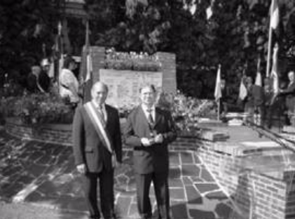 Two men in suits stand in front of a memorial with flowers and flags, whilst a group of people stands in the background.