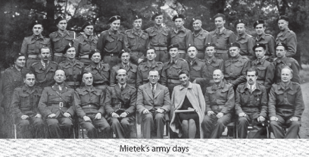 Black and white photo of a large group of soldiers in uniform, posed in three rows outdoors with four civilians seated in front.