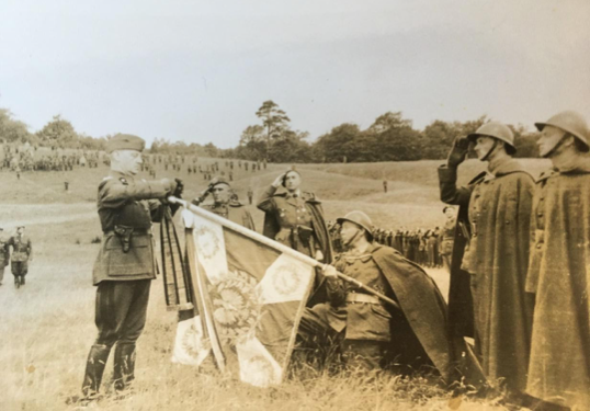 A military officer presents a flag to kneeling soldiers, whilst others stand to attention and salute on a grassy field.