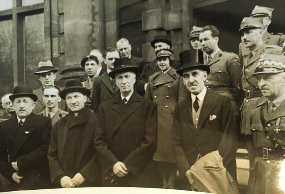 A group of men, some in military uniforms and others in formal suits and hats, pose for a photograph on the steps of a large stone building.
