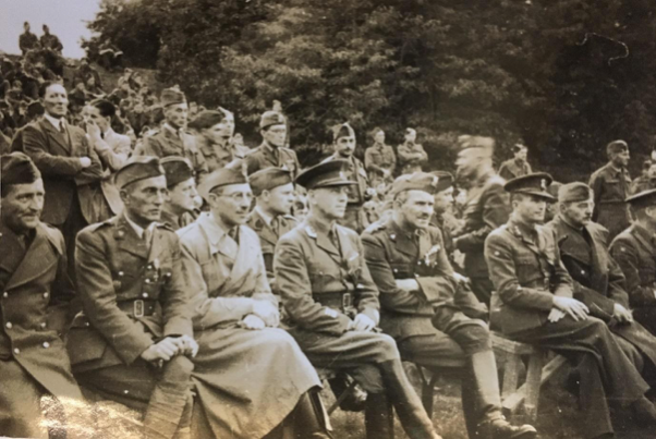 A group of military officers in uniform are seated outdoors in rows on benches, with trees in the background.