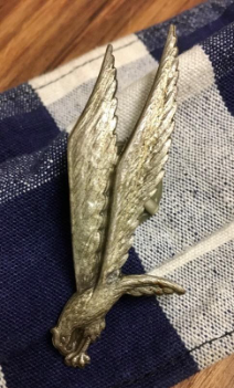 A metallic eagle-shaped pin with outstretched wings rests on a blue and white checked fabric.