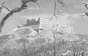 A damaged hilltop monastery, with ruined walls visible, stands behind leafless trees under a cloudy sky.