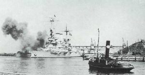A large battleship fires its guns near a harbour while a smaller vessel is seen in the foreground, with industrial structures in the background.