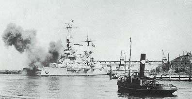 A large battleship fires its guns near a harbour while a smaller vessel is seen in the foreground, with industrial structures in the background.
