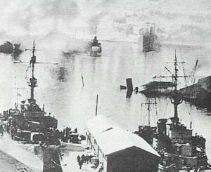 Black and white photo showing several warships docked in a harbour with smoke or haze in the background. The image appears historical and industrial.