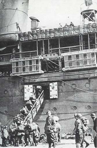 Soldiers in uniform board a large naval ship via a gangplank, whilst others stand and walk nearby on the dockside.